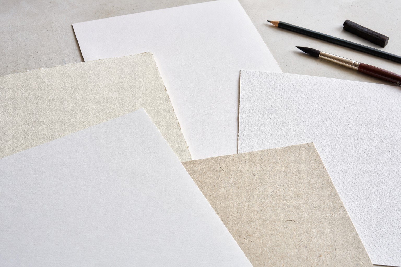 Vellen tekenpapier met verschillende korrels naast potlood, houtskool en penseel op een lichte werktafel